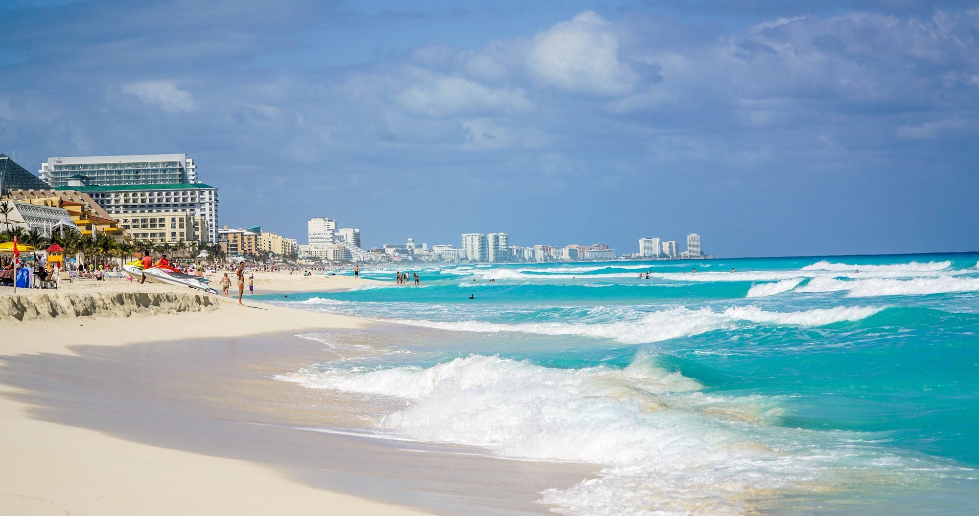 Ondas turquesa quebrando em uma praia de areia clara, com hotéis ao fundo, em Cancún.