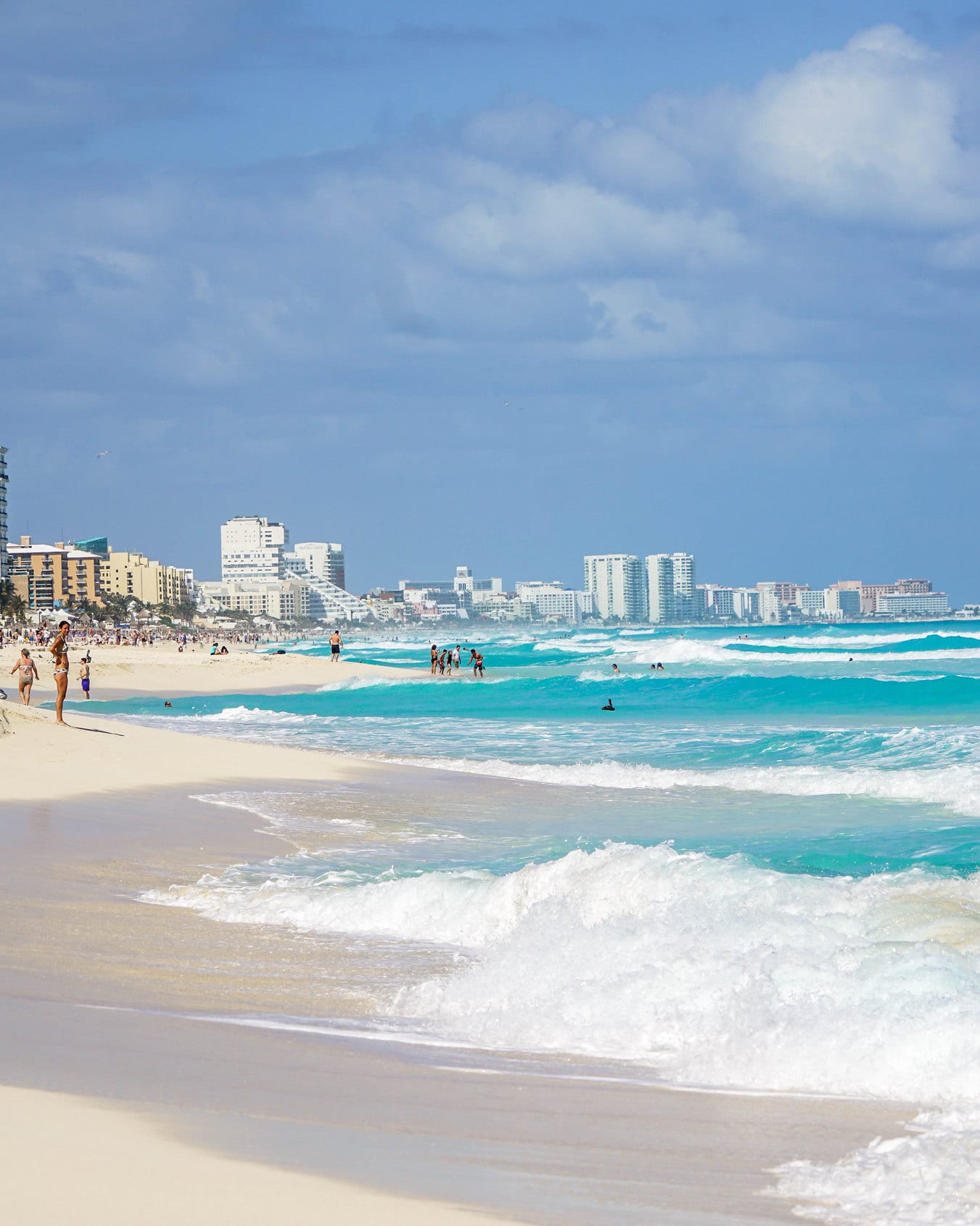 Ondas turquesa quebrando em uma praia de areia clara, com hotéis ao fundo, em Cancún.
