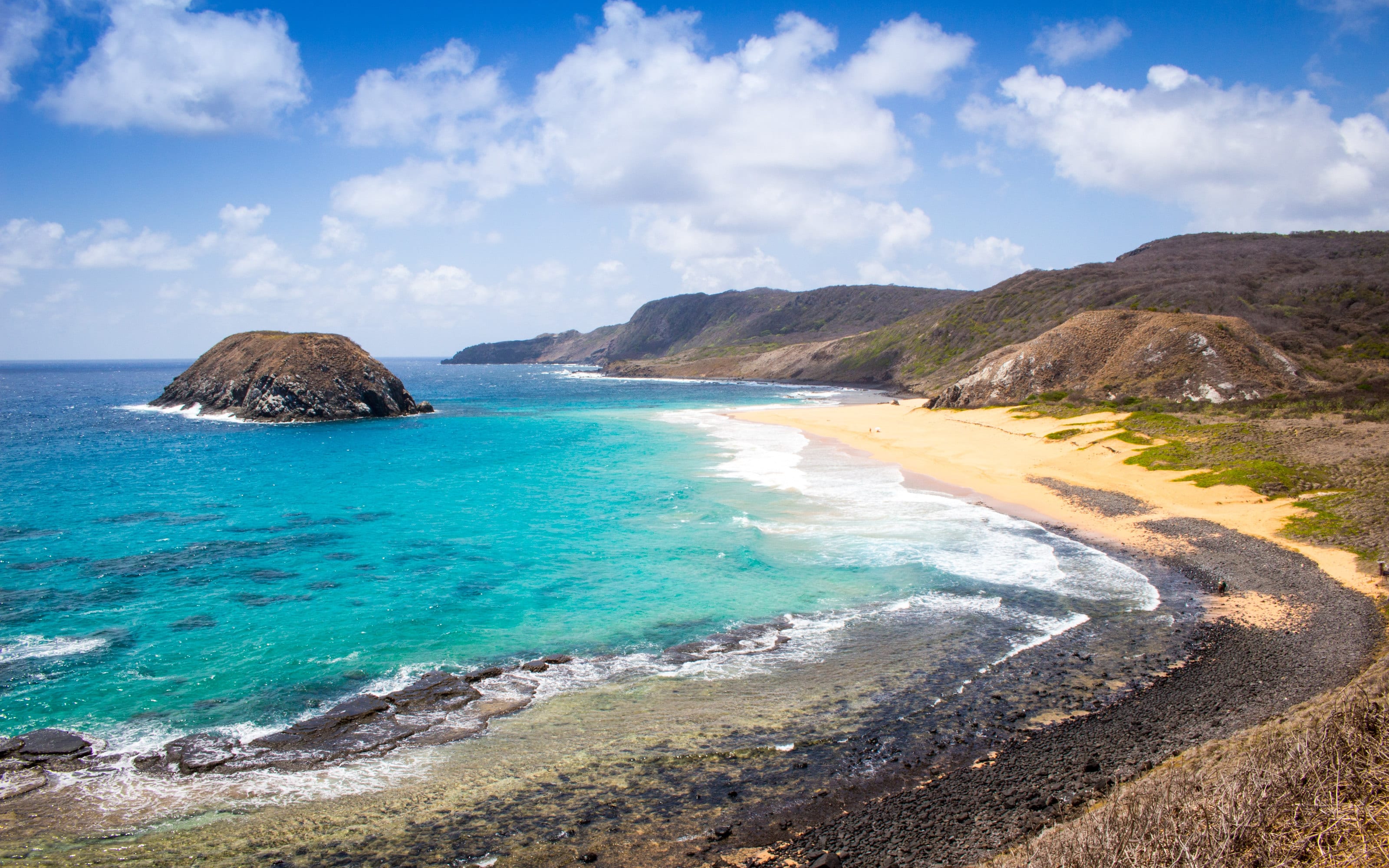 Praia do Leão, em Fernando de Noronha: mar turquesa de águas claras, faixa de areia dourada, falésias e um ilhéu rochoso sob céu azul.